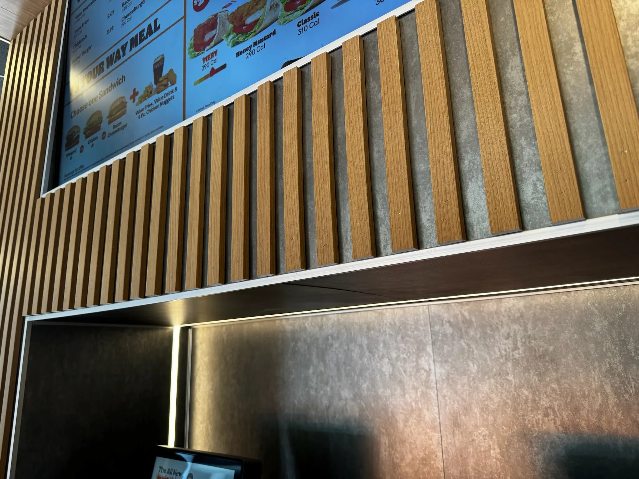 A close-up of a wall with vertical wooden slats beneath a fast food restaurant menu board displaying meal options, prices, and images of burgers and fries. A digital register screen is partially visible at the bottom.
