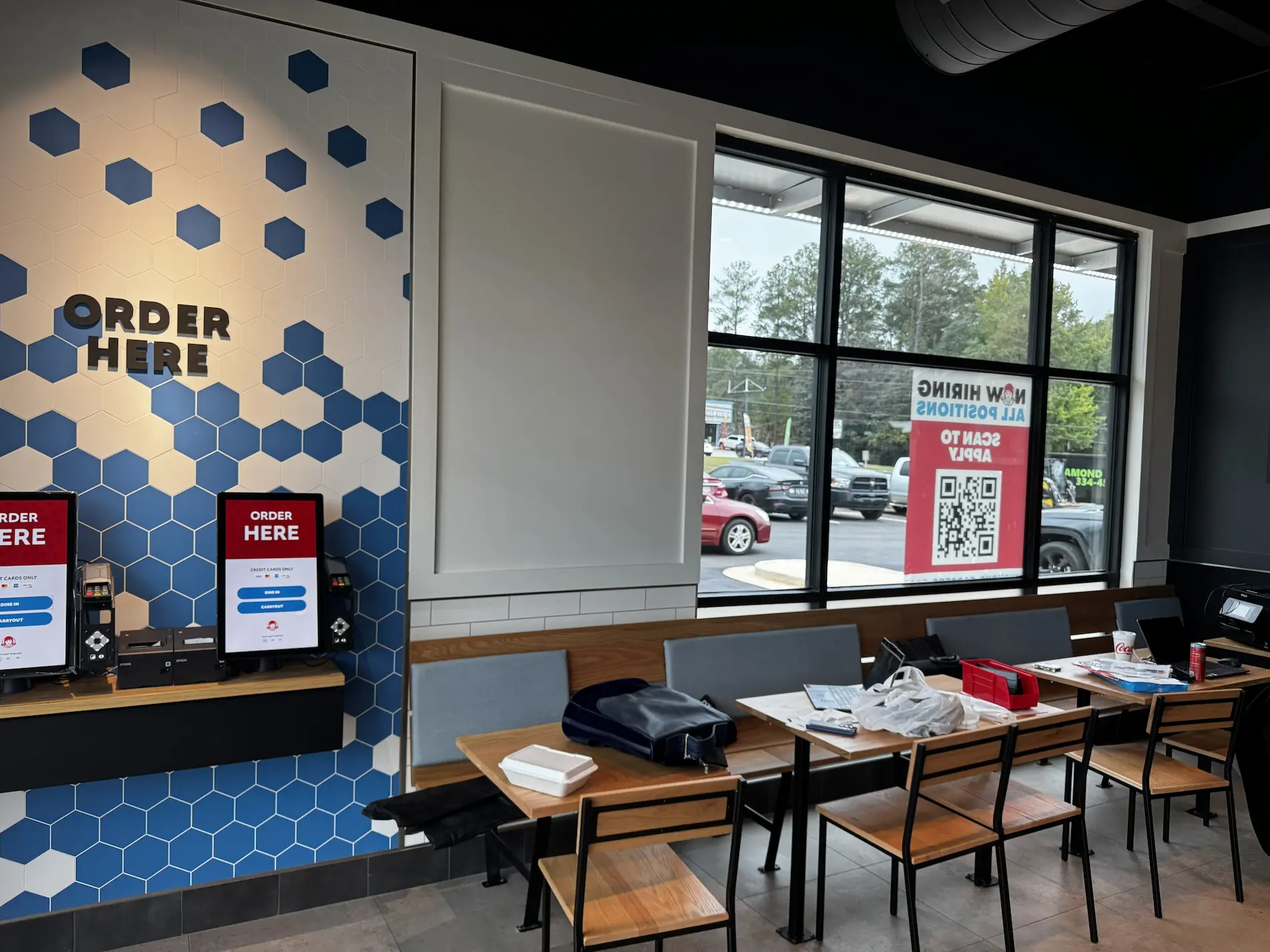 A fast food restaurant interior with self-order kiosks on a blue hexagon-patterned wall, empty tables and chairs, and a large window showing a “Now Hiring” sign and cars outside.