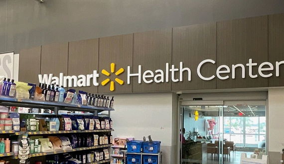 A Walmart Health Center sign is mounted on a wooden wall above the entrance, with shelves of health and beauty products in the foreground. The entrance features sliding glass doors.