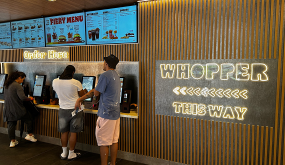 Three people use self-service kiosks under a menu board in a fast-food restaurant. A neon sign on the wall says WHOPPER THIS WAY with arrows pointing right.