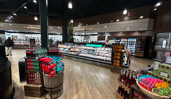 A grocery store interior with wide aisles, refrigerated display cases of meats and dairy, baskets of snacks, and shelves of drinks. Shoppers and a Christmas tree are visible in the background.