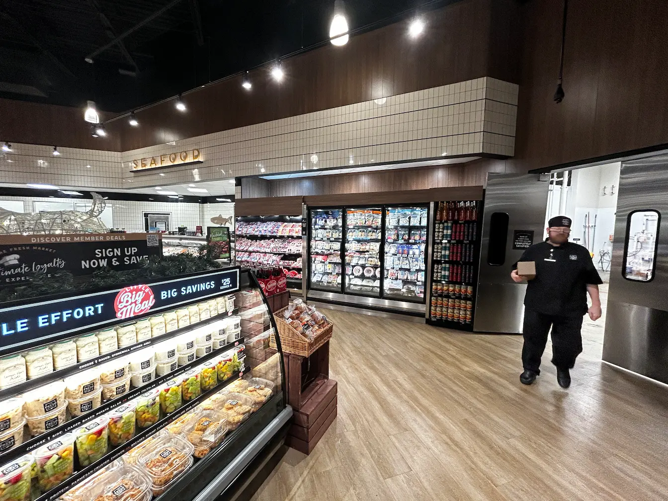 A grocery store interior shows refrigerated seafood section, prepared foods, grocery displays, and a worker in uniform walking through a doorway. The floor is light wood, and the lighting is bright and modern.